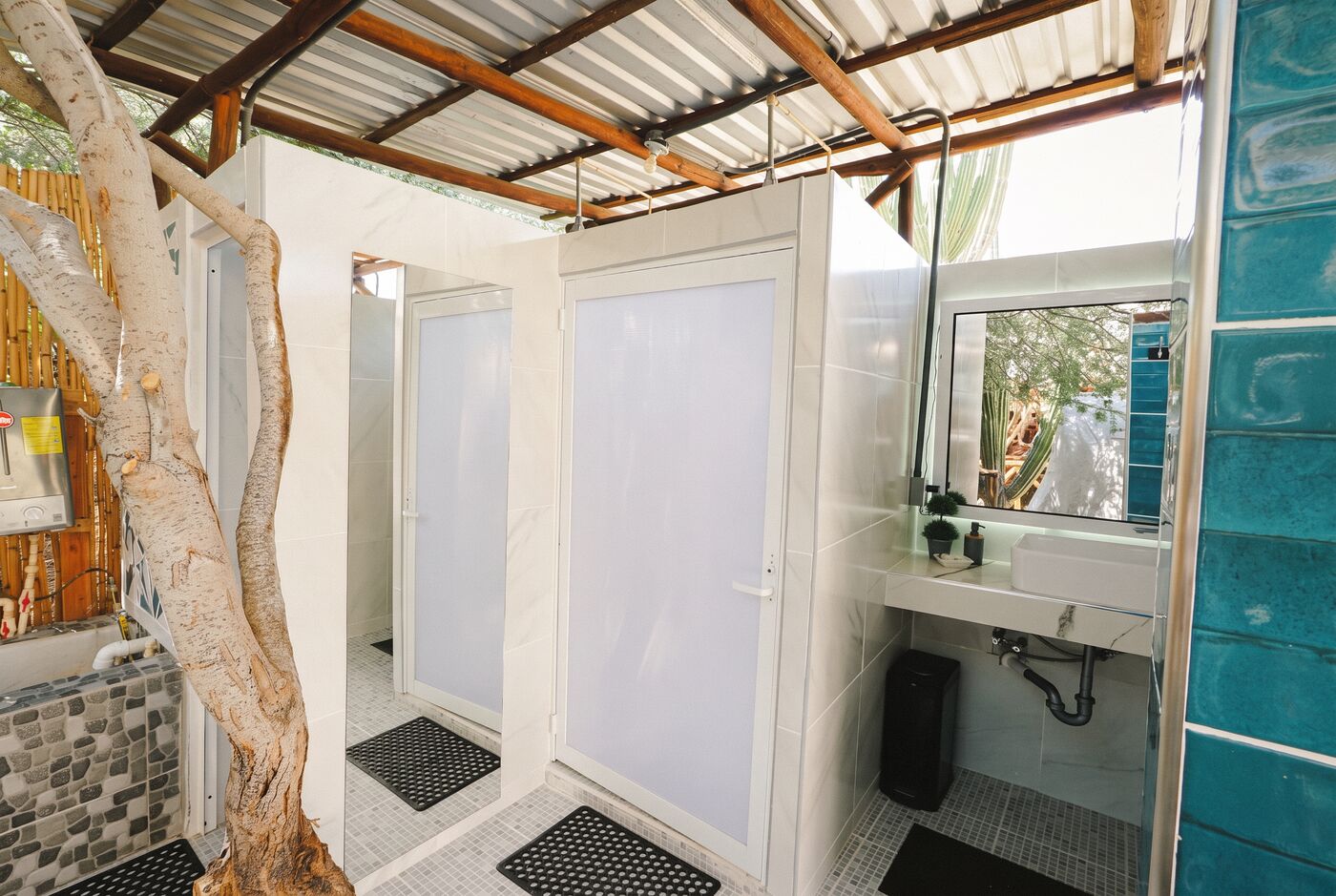 The shared bathrooms of the glamping tents — white-tiled cabins, a turquoise tiled wall and a living tree trunk