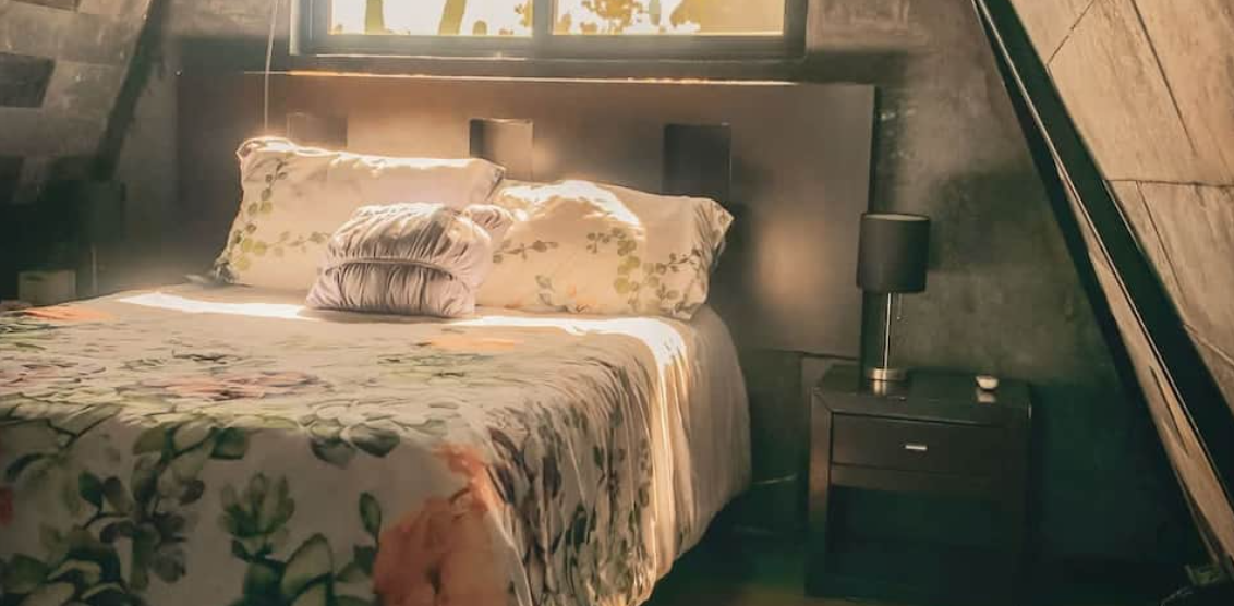 Tapanco Suite — bed with botanical-print linens under a sloped wall, warm light from a high window